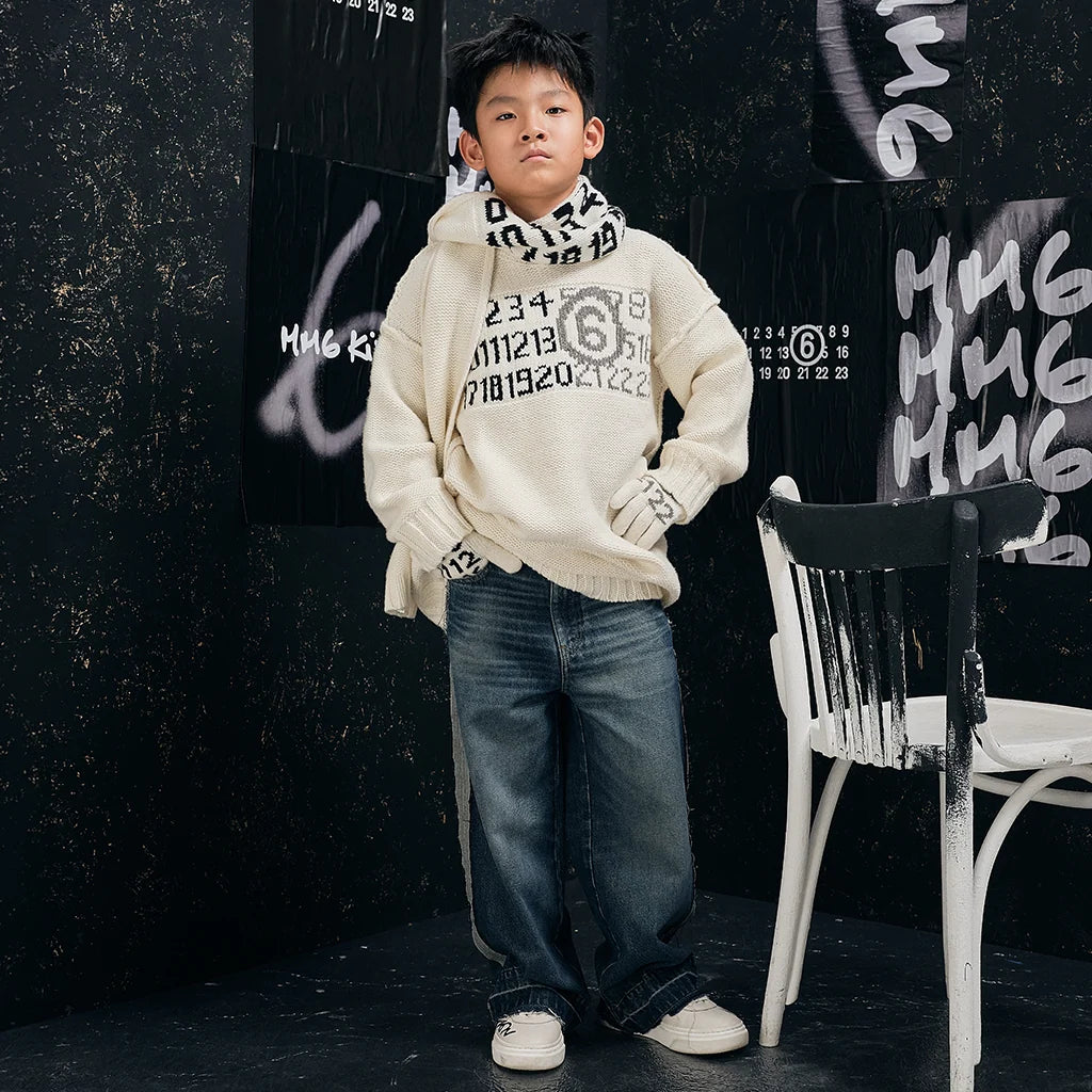 Boy standing against a black background, wearing a stylish and warm look composed of a pullover designed by MM6 Maison Margiela. 