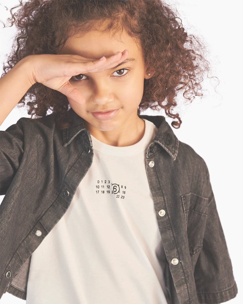 Child wearing a dark denim jacket over a white shirt with a logo, posing with hand on forehead.